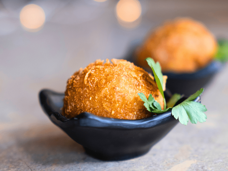 "Croqueta dorada y crujiente servida en cuenco negro con decoración de perejil fresco y bokeh de luces al fondo"