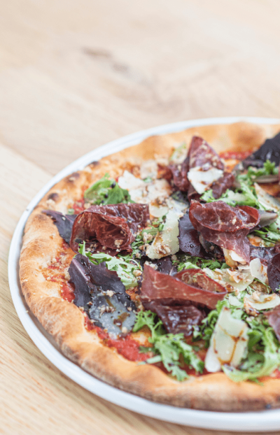 "Pizza artesana con masa fina y crujiente, topped con bresaola, rúcula fresca y láminas de parmesano sobre base de tomate"