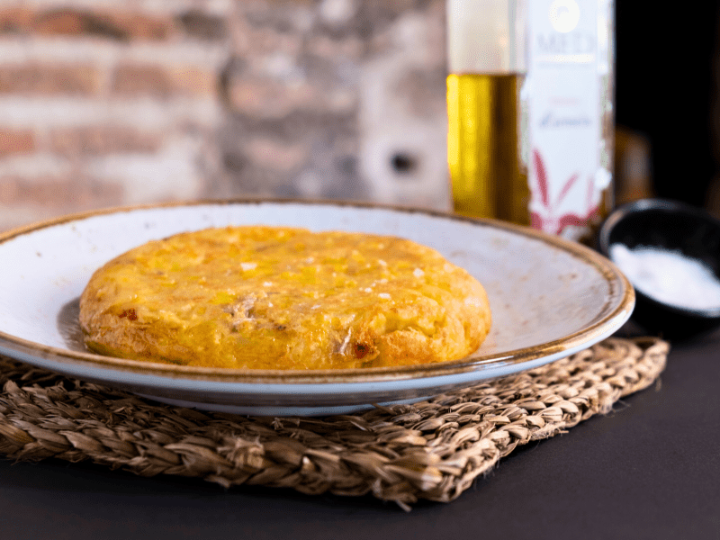 "Tortilla española dorada y jugosa servida en plato blanco rústico sobre salvamanteles de fibra natural"
